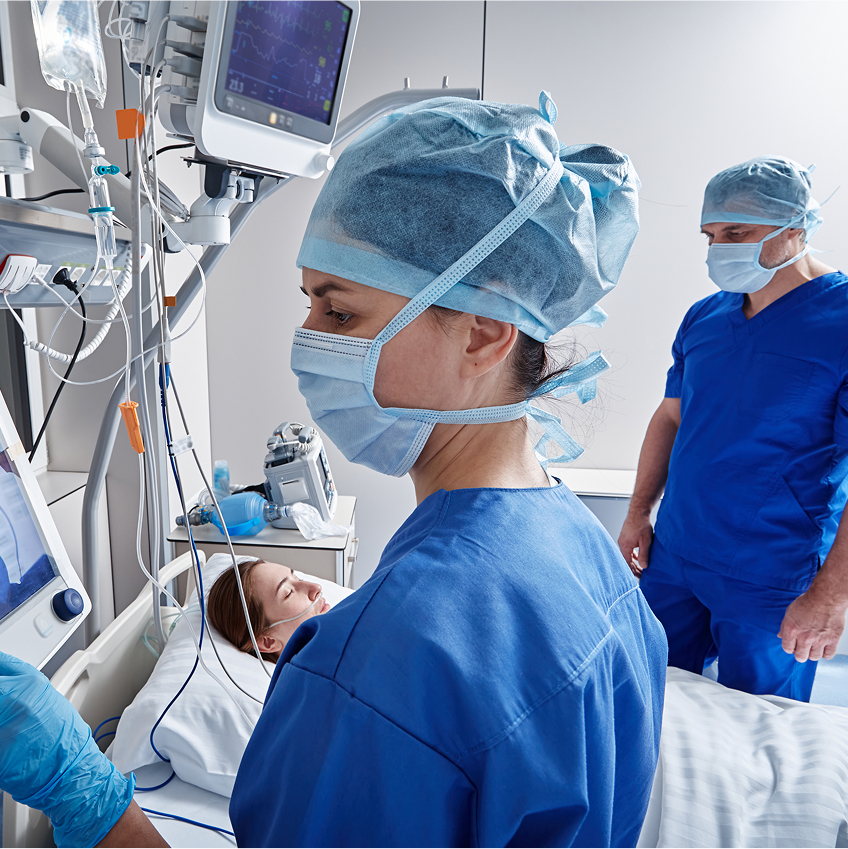 Zwei medizinische Fachkräfte in blauer Kleidung und Masken stehen neben einem Patienten im Krankenhausbett.