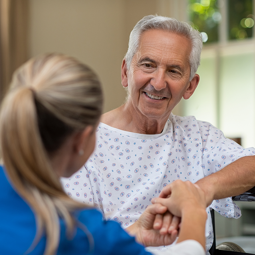 Ein älterer Mann in einem Krankenhaushemd lächelt und spricht mit einer Krankenschwester, die ihm gegenüber sitzt.