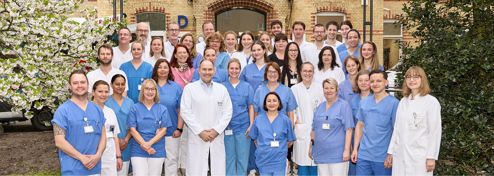 Gruppe von medizinischem Personal in blauer und weißer Arbeitskleidung posiert im Außenbereich der Klinik für ein Foto.