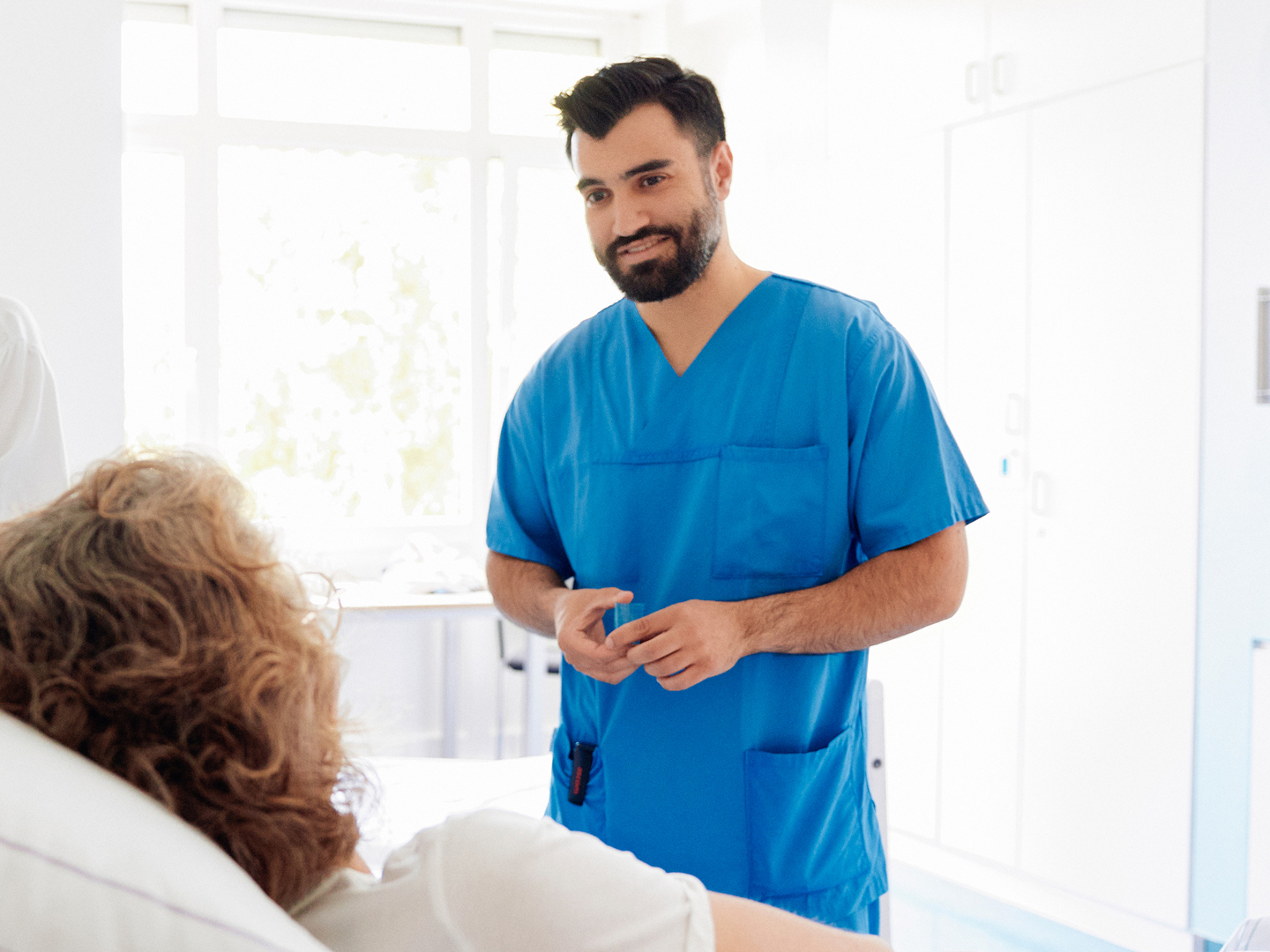 Eine medizinische Fachkraft in blauer Arbeitskleidung spricht freundlich mit einer Patientin, die im Krankenhausbett liegt.