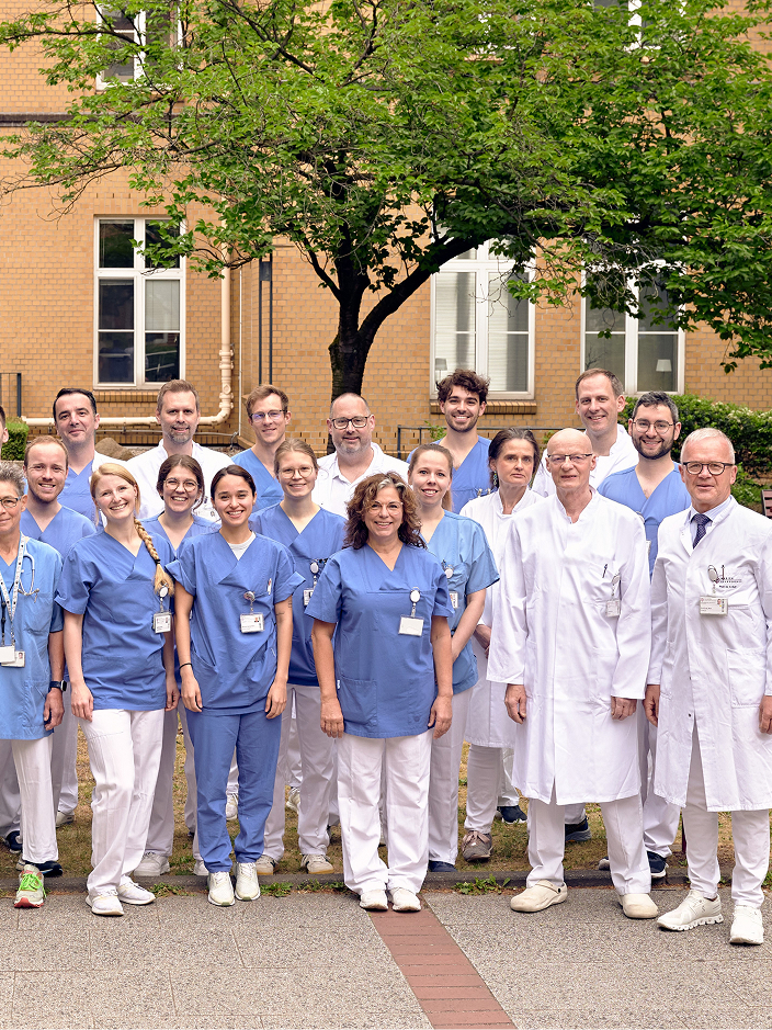 Gruppe von 20 medizinischen Fachkräften in blauen und weißen Kitteln steht vor dem Klinikgebäude und posiert für ein Foto.