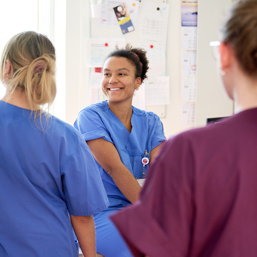 Eine lächelnde Krankenschwester in blauer Uniform sitzt, umgeben von zwei weiteren Personen im Stationszimmer.