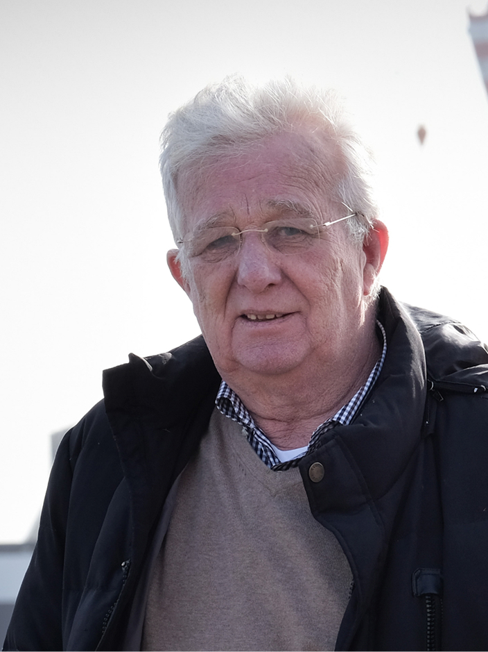 Älterer Mann mit Brille, grauen Haaren und dunkler Jacke, steht im Freien vor einem hellen Hintergrund.