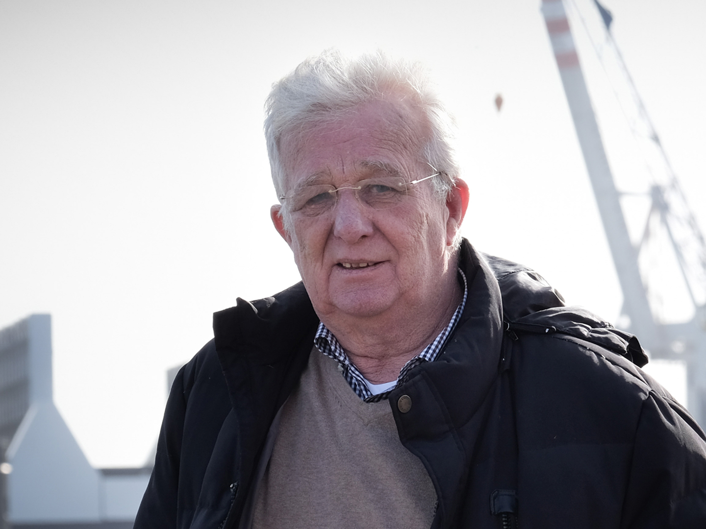 Älterer Mann mit Brille und grauen Haaren, trägt eine dunkle Jacke, er steht im Freien vor einem unscharfen Hintergrund.