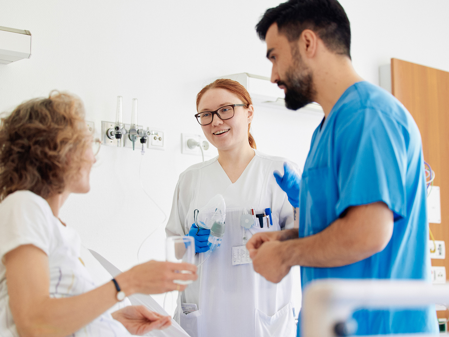Eine Patientin im Krankenhausbett hält ein Glas Wasser, während sie mit zwei medizinischen Fachkräften im Gespräch ist.