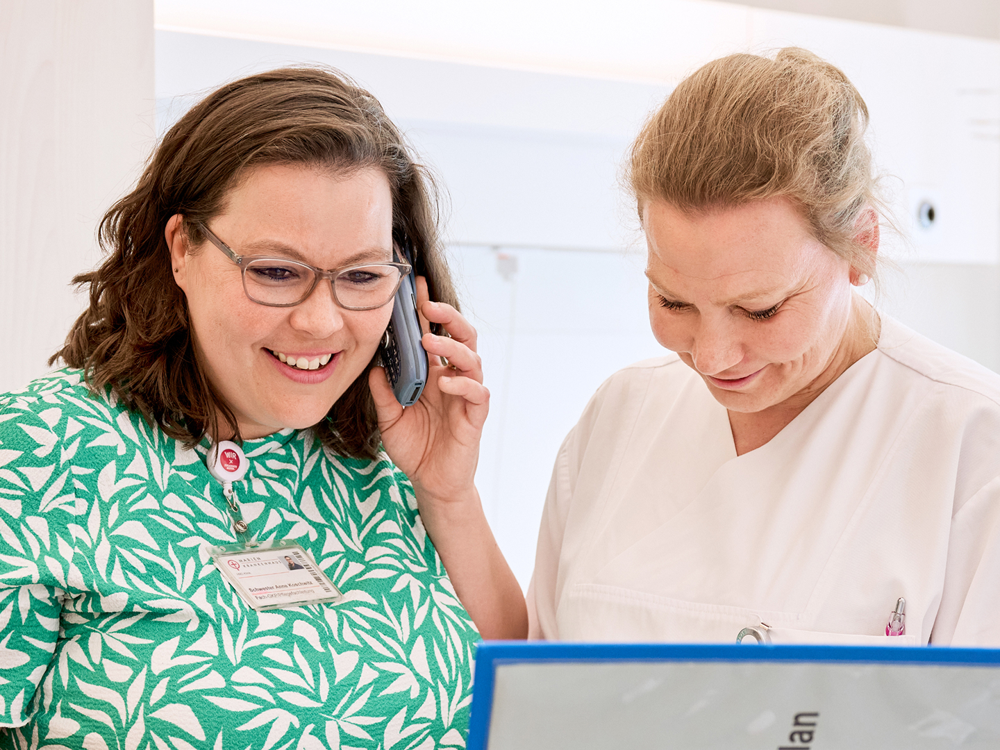 Zwei Frauen stehen nebeneinander in einem hellen Raum, eine telefoniert und die andere schaut in einen Ordner.