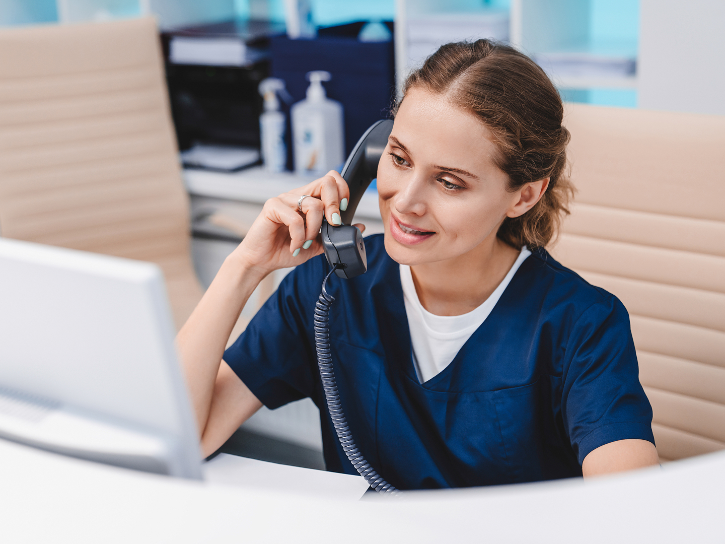 Frau in blauer Arbeitskleidung spricht am Telefon, während sie an einem Schreibtisch mit Computer sitzt.