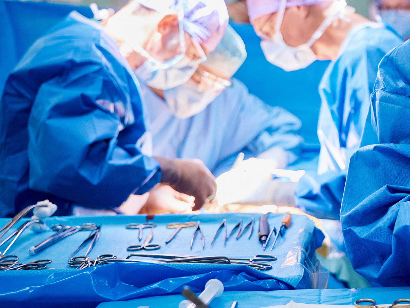 Chirurgisches Team in blauen Kitteln führt eine Operation durch, im Vordergrund sieht man chirurgische Instrumente.