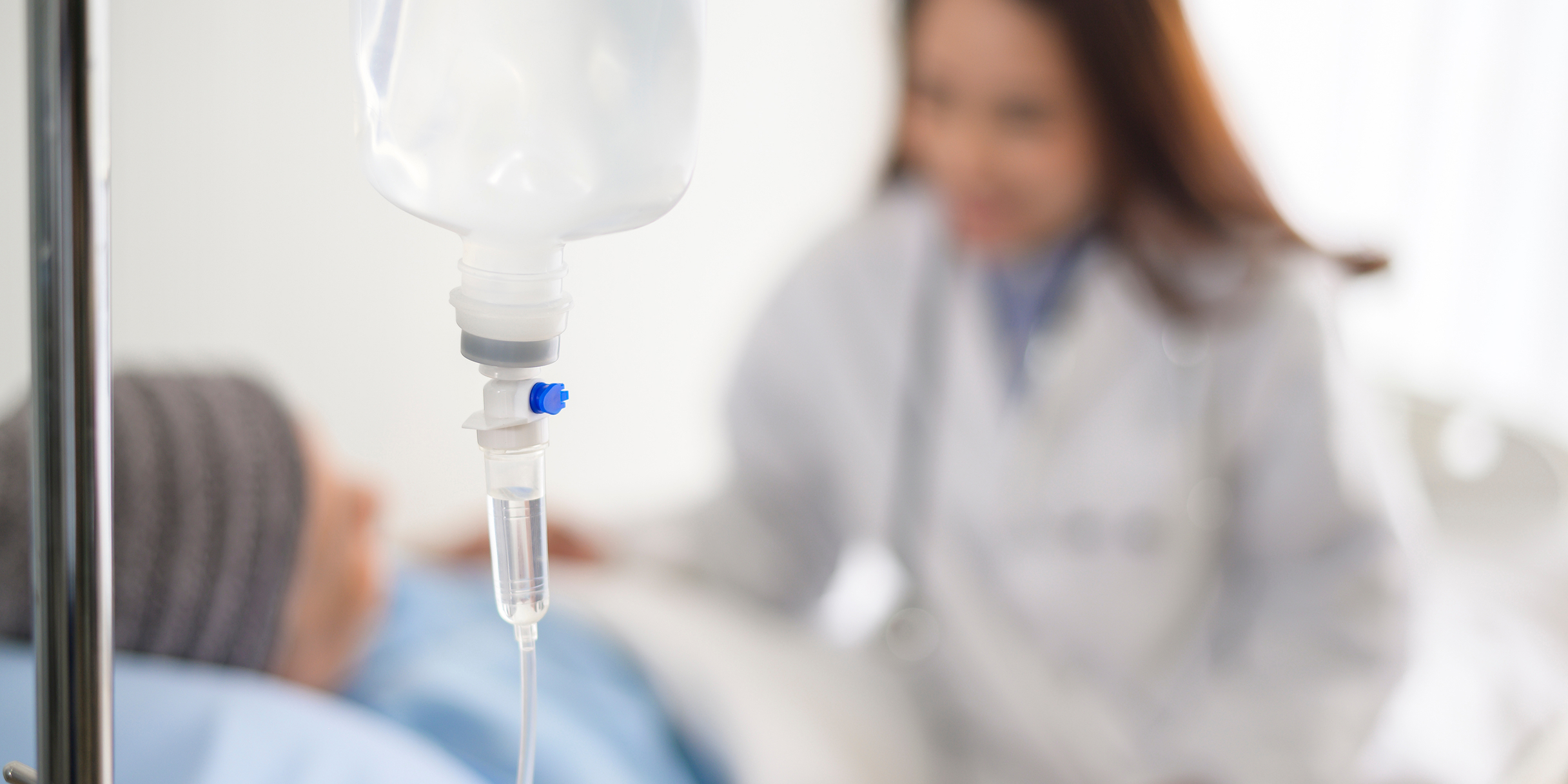 Infusionsbeutel mit Tropfleitung im Vordergrund, im Hintergrund eine Ärztin, die sich um einen Patienten kümmert.