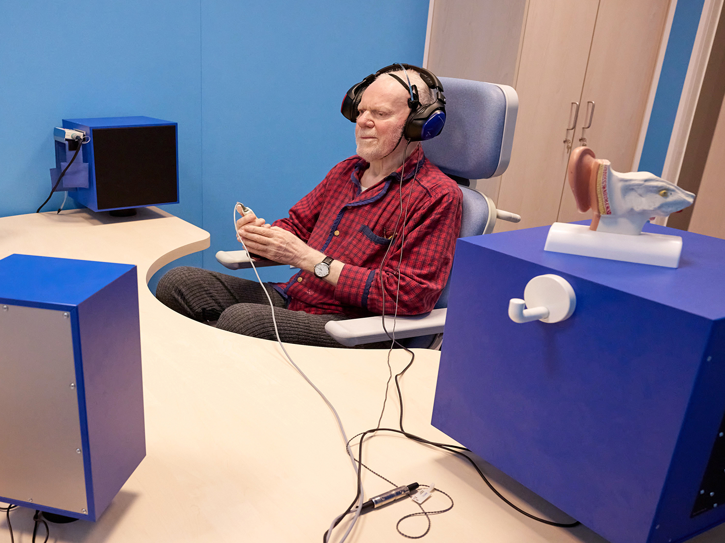 Ein älterer Mann sitzt in einem Stuhl, trägt Kopfhörer und hält ein Gerät, das mit einer blauen Box und einem Monitor verbunden ist.