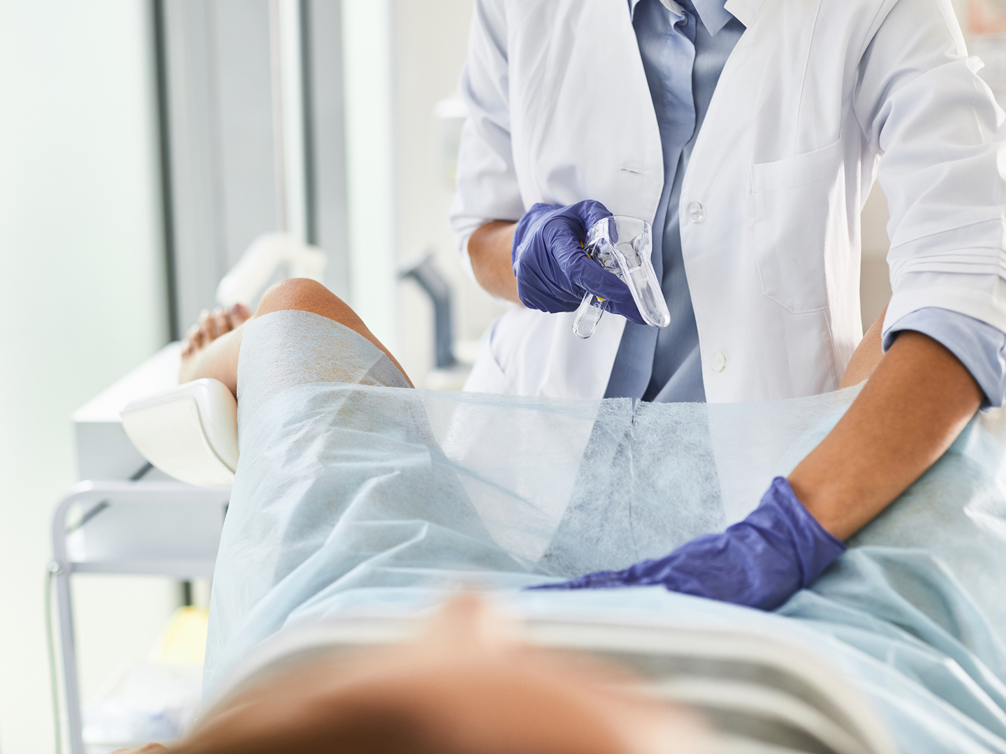 Eine medizinische Fachkraft in einem weißen Kittel bereitet eine gynäkologische Untersuchung bei einer Patientin vor, die auf einer Liege liegt.