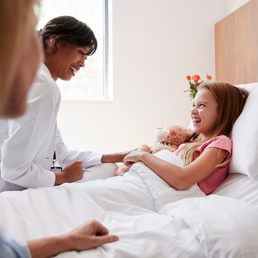 Ärztin spricht lächelnd mit einem Mädchen im Bett, das ein Stofftier hält, während eine weitere Person zuschaut.
