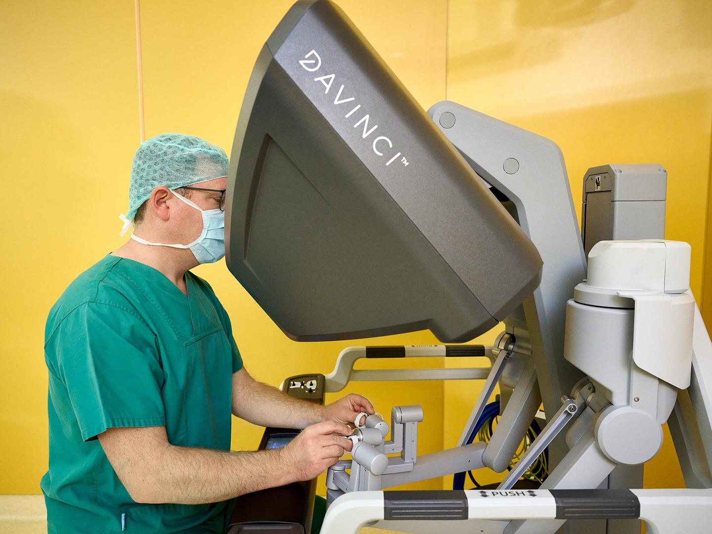 Ein Arzt in grüner OP-Kleidung bedient ein Da Vinci-Operationssystem in einem gelben Raum.