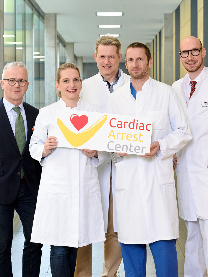 Fünf Personen in weißen Kitteln stehen im Klinikflur, zwei davon halten ein Schild mit der Aufschrift 