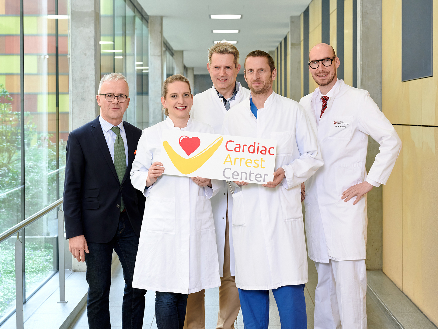 Fünf Personen in weißen Kitteln stehen im Klinikflur, zwei davon halten ein Schild mit der Aufschrift 