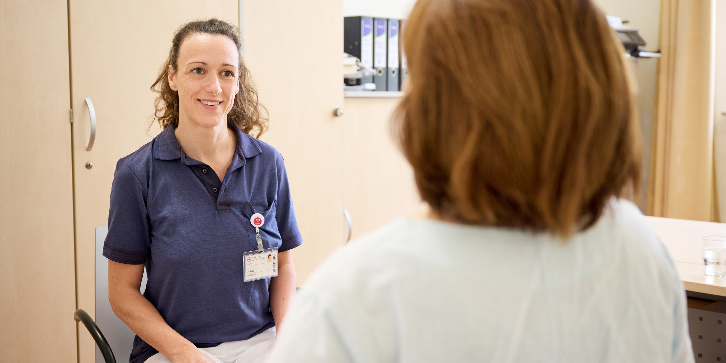 Eine Krankenschwester in blauer Arbeitskleidung spricht mit einer Patientin in einem Besprechungsraum.