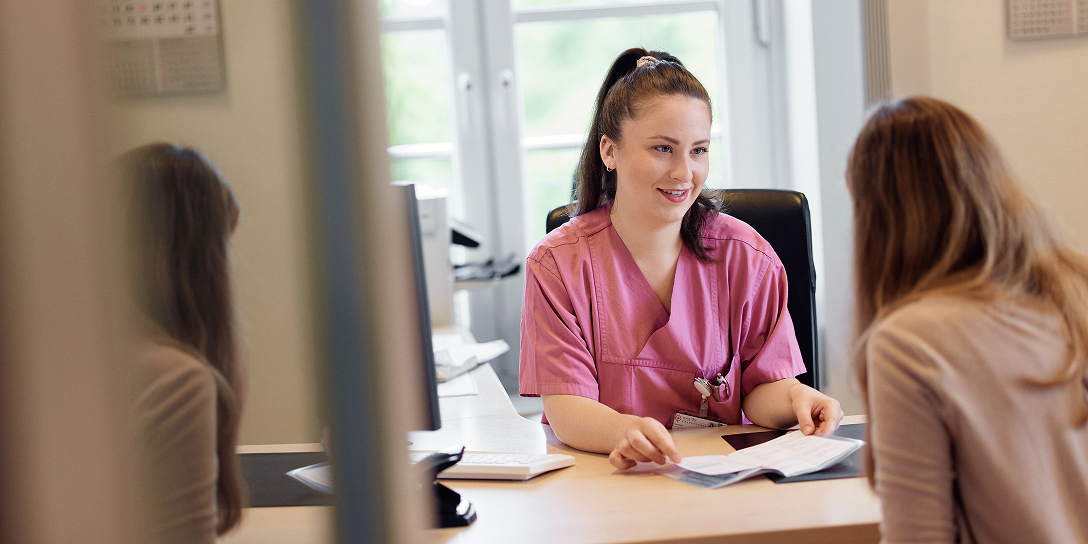 Eine lächelnde medizinische Fachkraft in rosafarbener Arbeitskleidung sitzt an einem Schreibtisch und spricht mit einer Patientin.