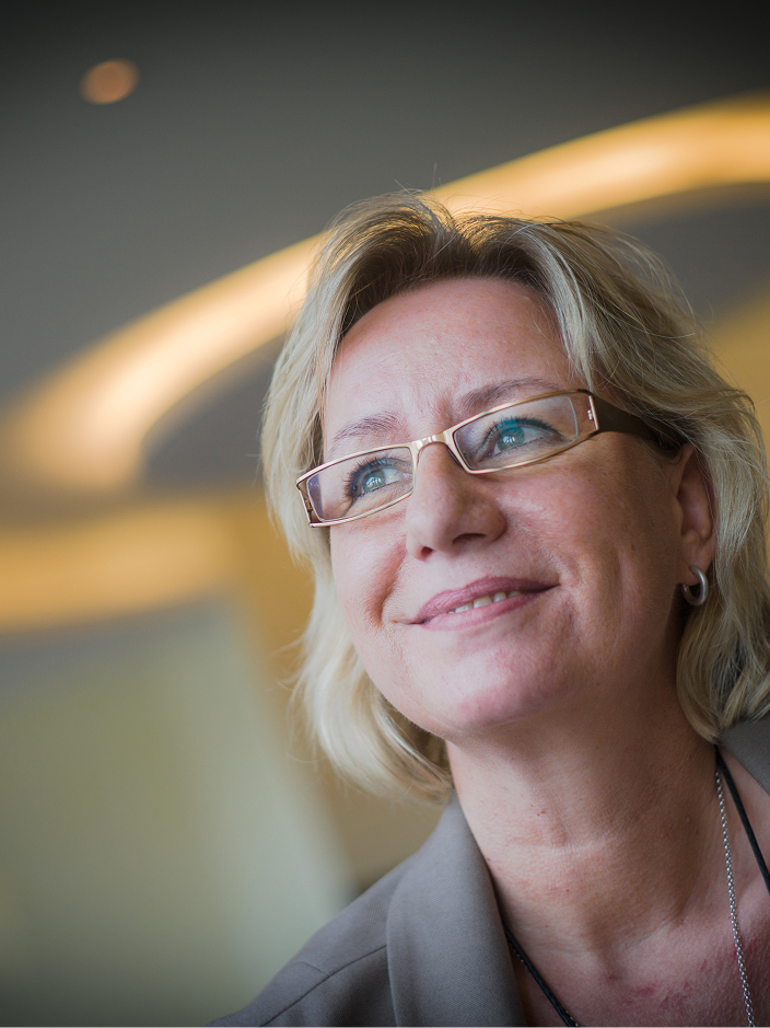 Frau mit blonden Haaren und Brille lächelt, im Hintergrund sanftes Licht und unscharfe Formen.