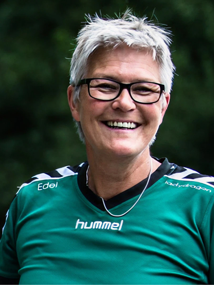 Lächelnde Person mit kurzen grauen Haaren, Brille und grünem Sporttrikot, vor unscharfem Hintergrund.