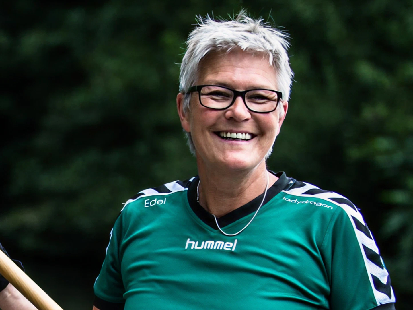 Lächelnde Person mit kurzen grauen Haaren, Brille und grünem Sporttrikot, vor unscharfem Hintergrund.