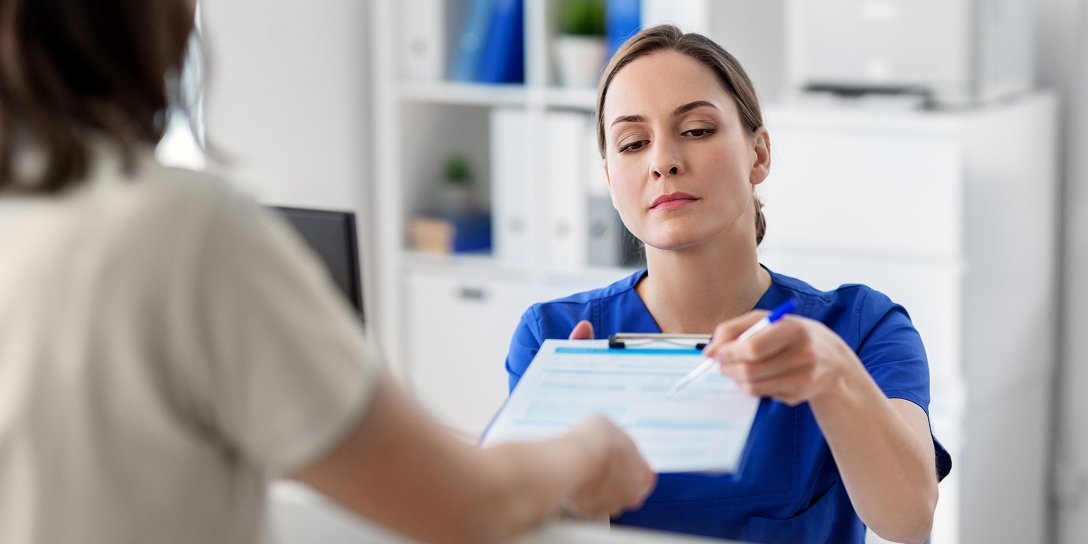 Eine Frau in blauer Arbeitskleidung reicht einer anderen Person ein Formular über den Empfangstresen hinweg. Sie hält einen Stift in der Hand.