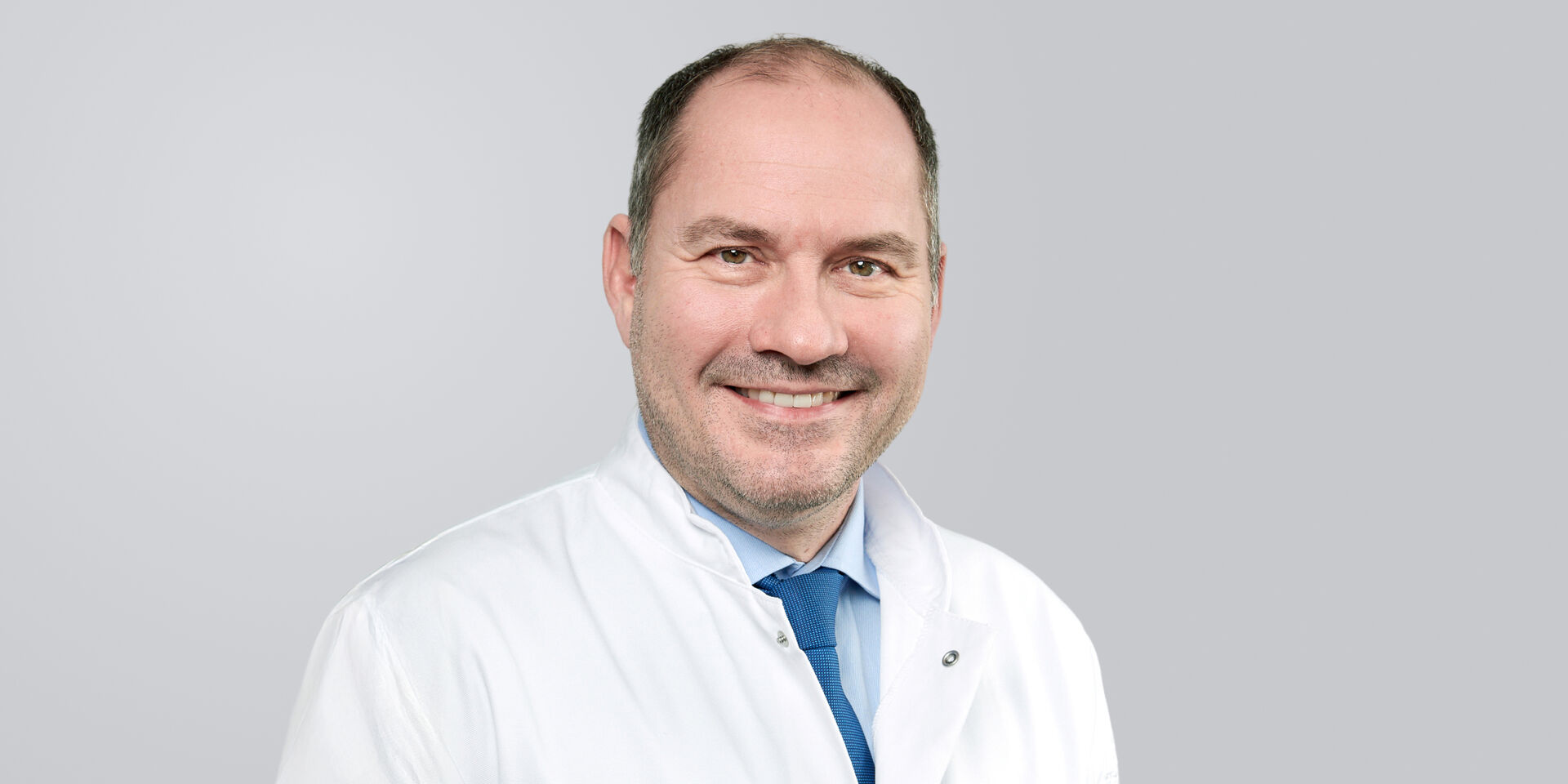 Lächelnder Mann mit Dreitagebart, weißem Kittel und blauer Krawatte vor grauem Hintergrund.