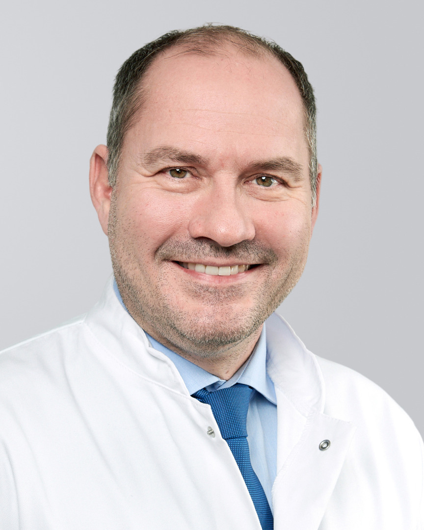 Lächelnder Mann mit Dreitagebart, weißem Kittel und blauer Krawatte vor grauem Hintergrund.