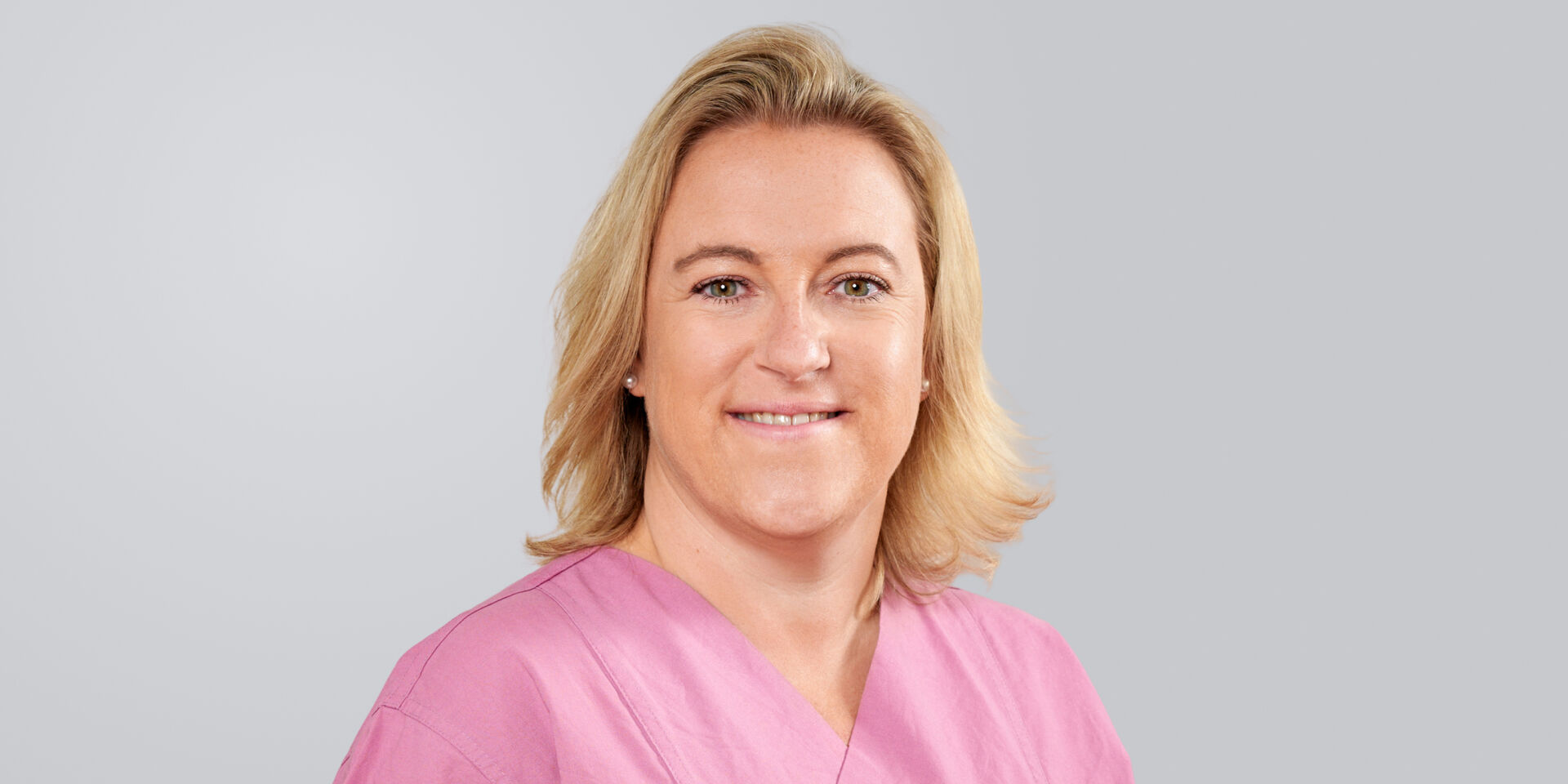 Frau mit schulterlangen, blonden Haaren in einem rosafarbenen Oberteil lächelt vor einem grauen Hintergrund.