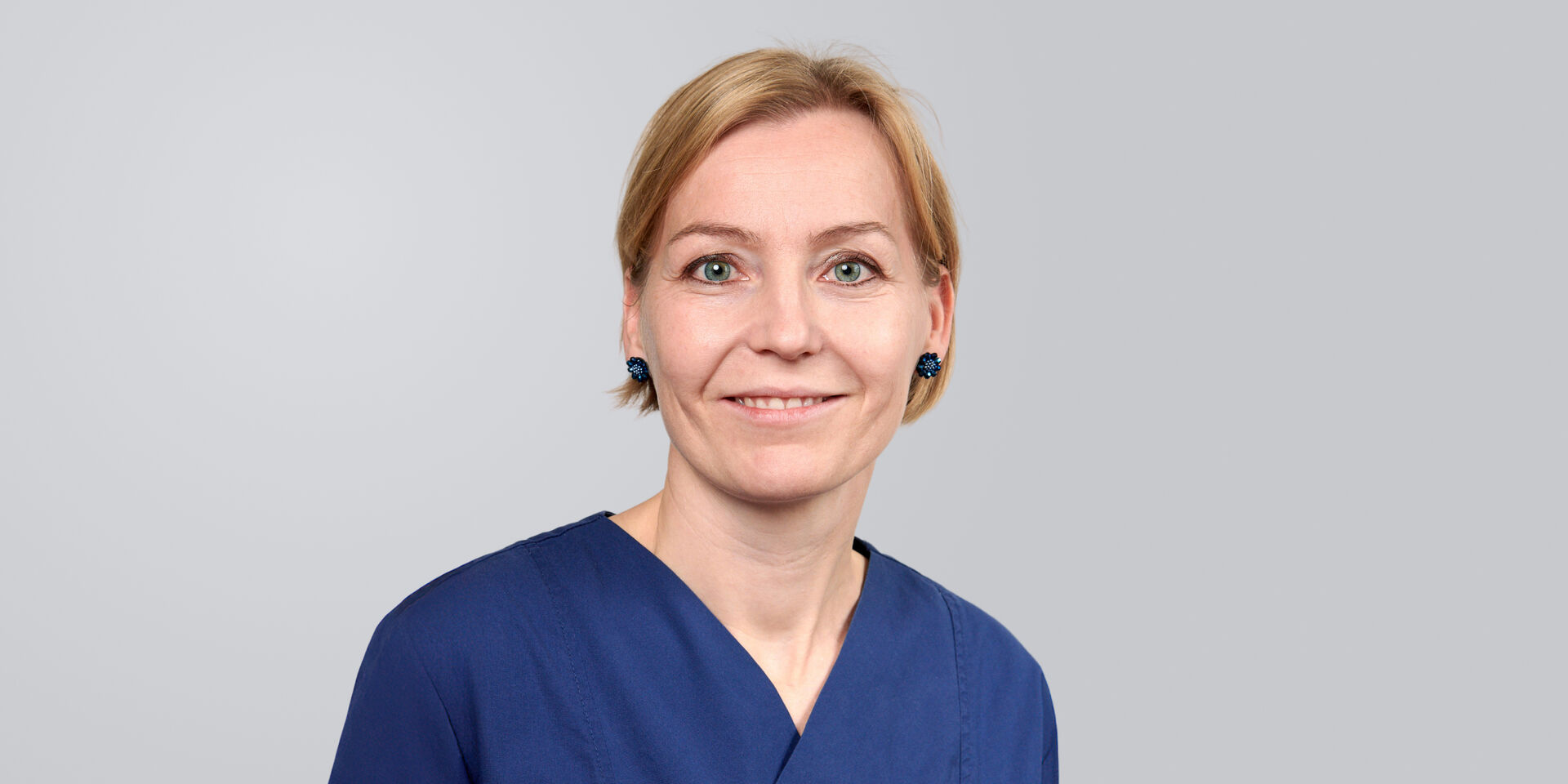 Frau mit kurzen, blonden Haaren in blauer Arbeitskleidung vor neutralem Hintergrund.