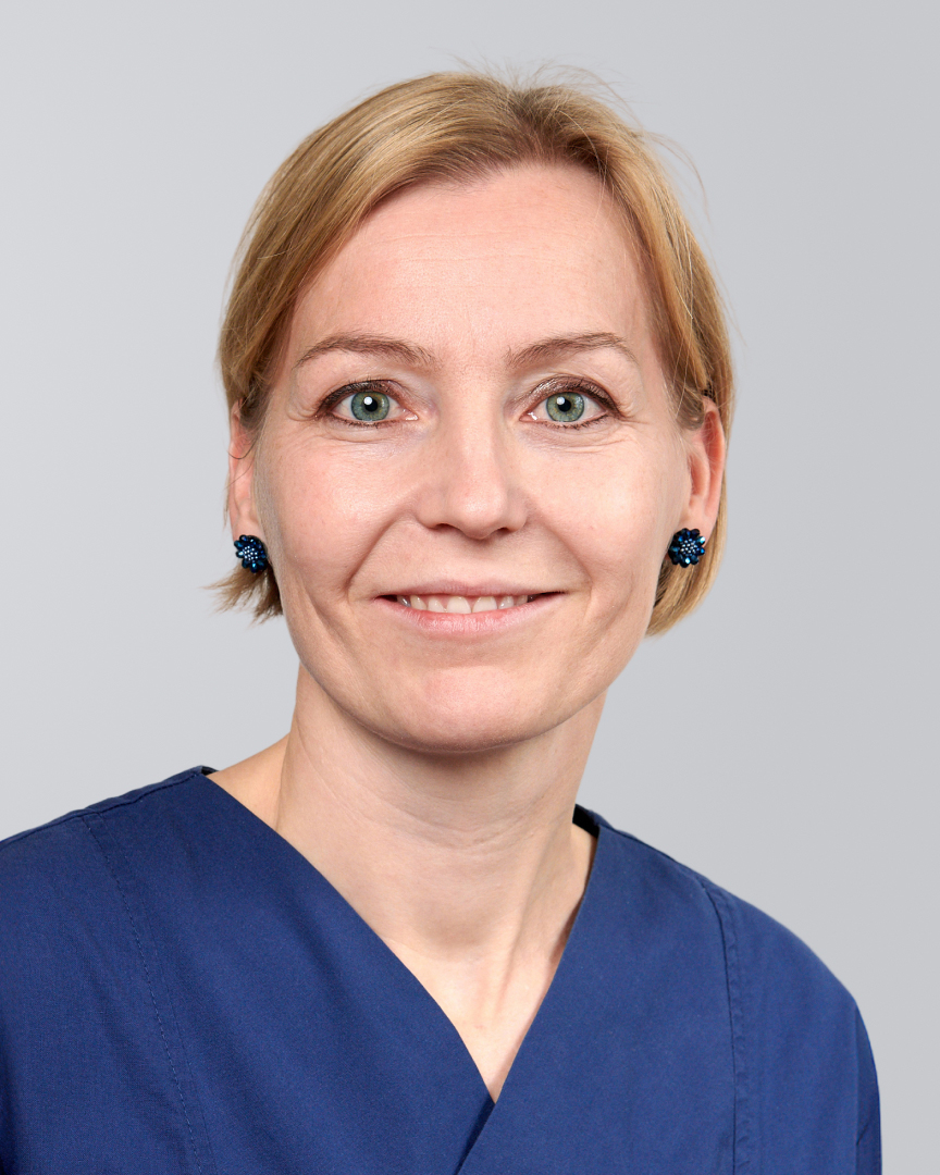Frau mit kurzen, blonden Haaren in blauer Arbeitskleidung vor neutralem Hintergrund.