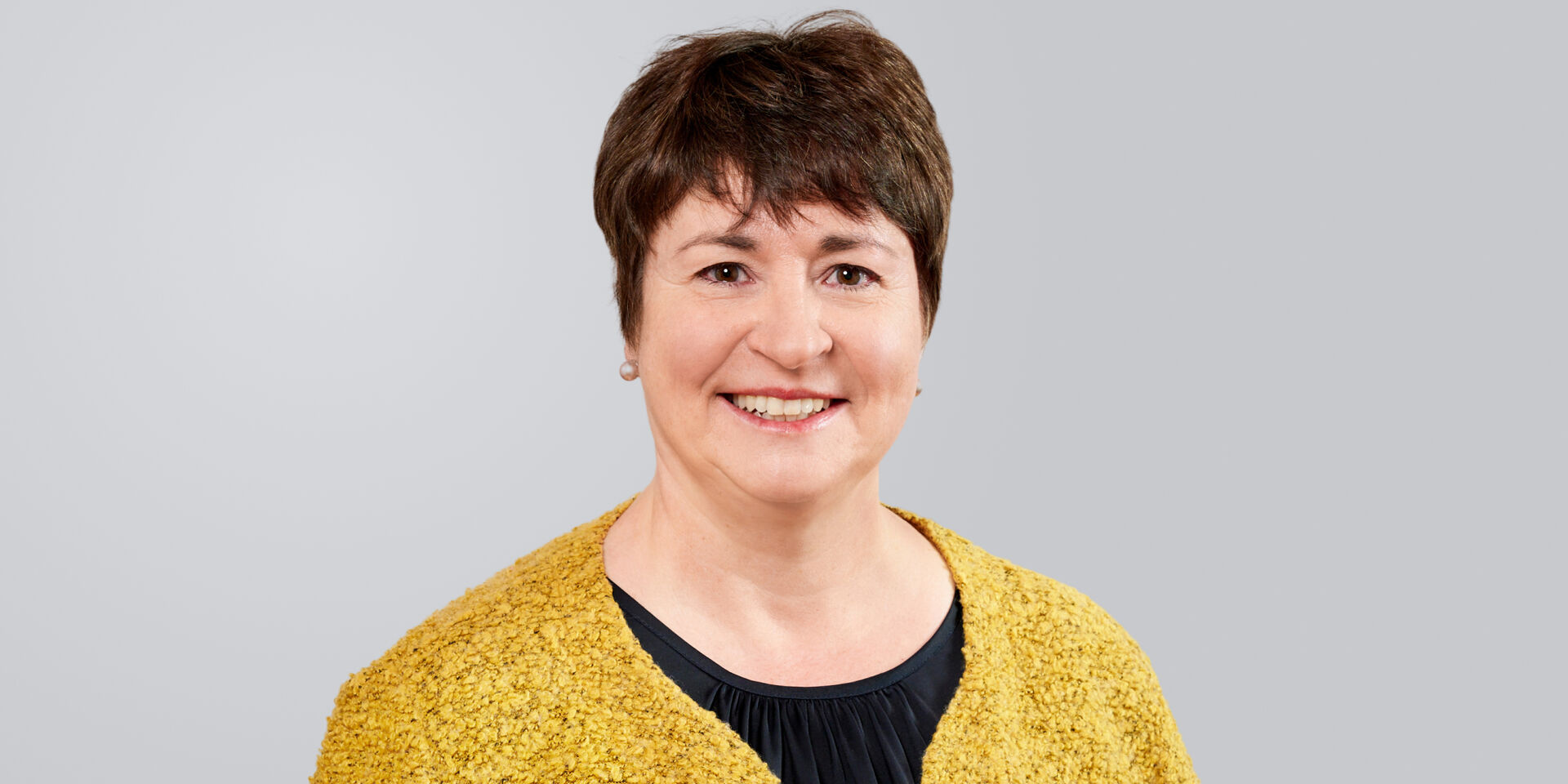 Porträt einer lächelnden Frau mit kurzen, braunen Haaren, die eine gelbe Strickjacke über einem schwarzen Oberteil trägt.