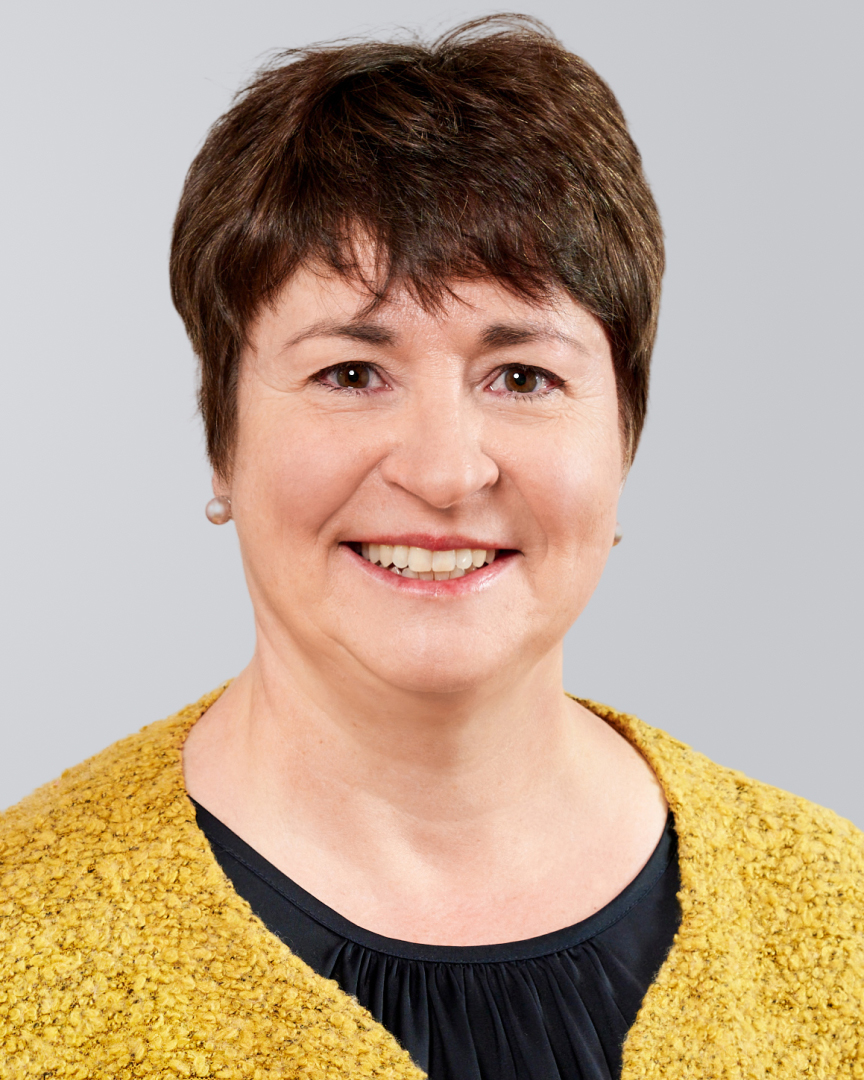 Porträt einer lächelnden Frau mit kurzen, braunen Haaren, die eine gelbe Strickjacke über einem schwarzen Oberteil trägt.