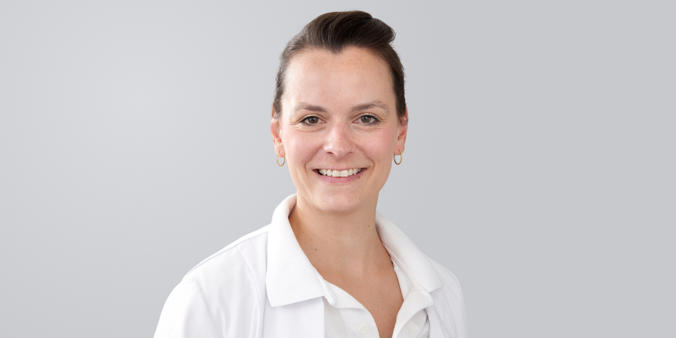 Lächelnde Frau mit dunklen Haaren und Arztkittel vor grauem Hintergrund.