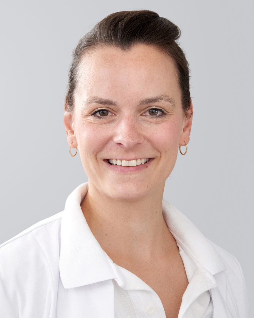 Lächelnde Frau mit dunklen Haaren und Arztkittel vor grauem Hintergrund.