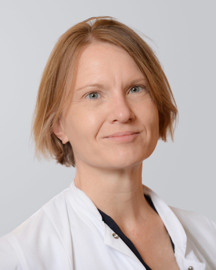Frau mit kurzen, dunkelblonden Haaren und weißen Kittel lächelt vor einem neutralen Hintergrund.