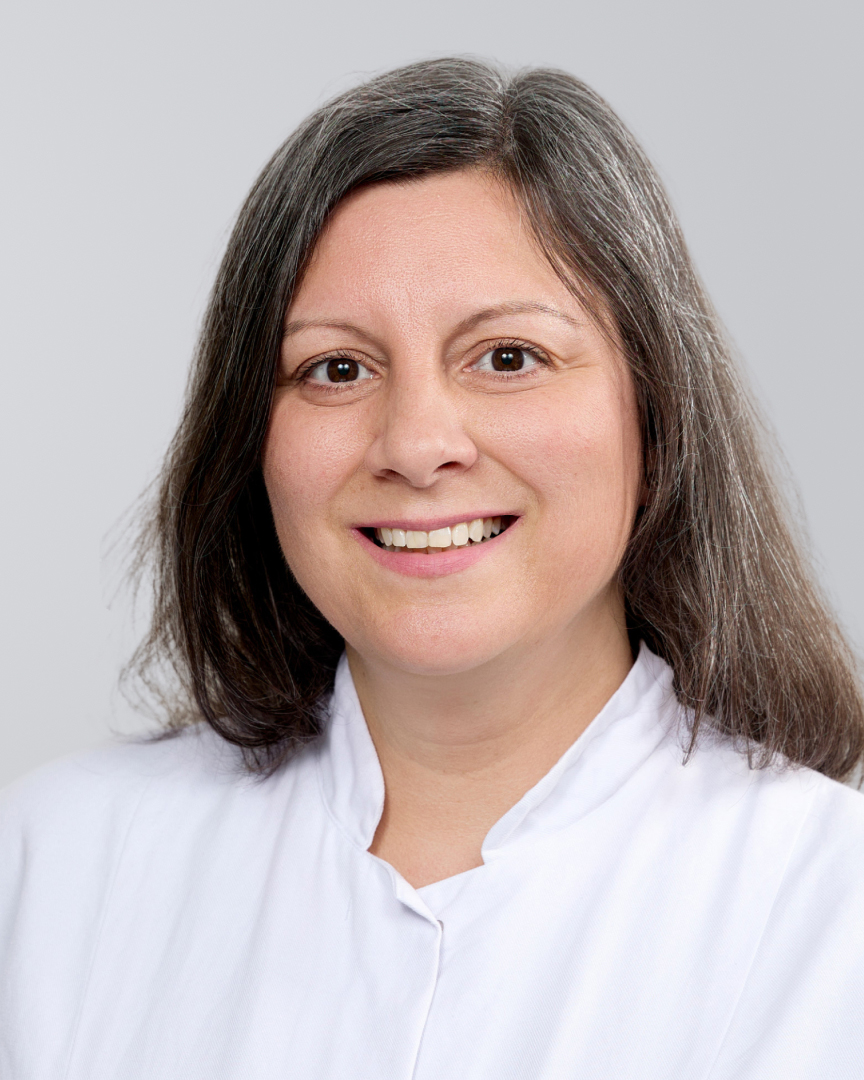 Frau mit langen, dunklen Haaren in einem weißen Kittel lächelt vor einem grauen Hintergrund.