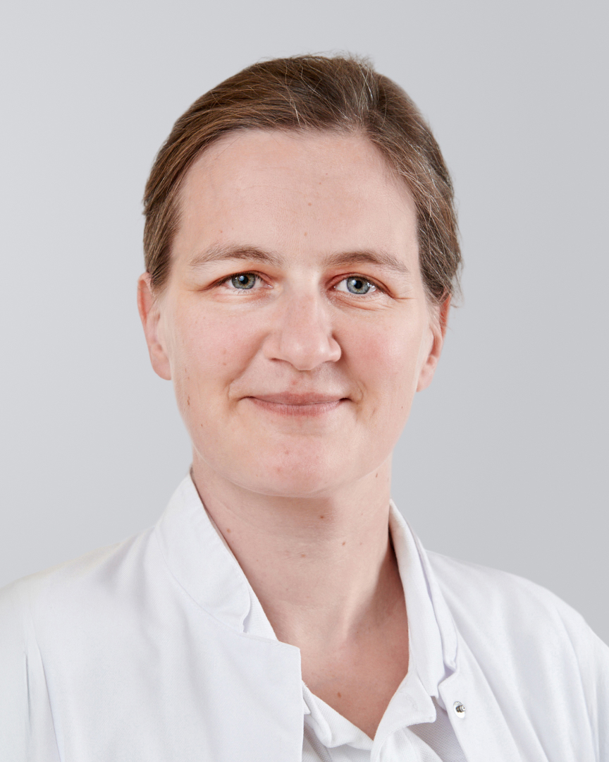 Frau in weißem Kittel lächelt vor neutralem Hintergrund.