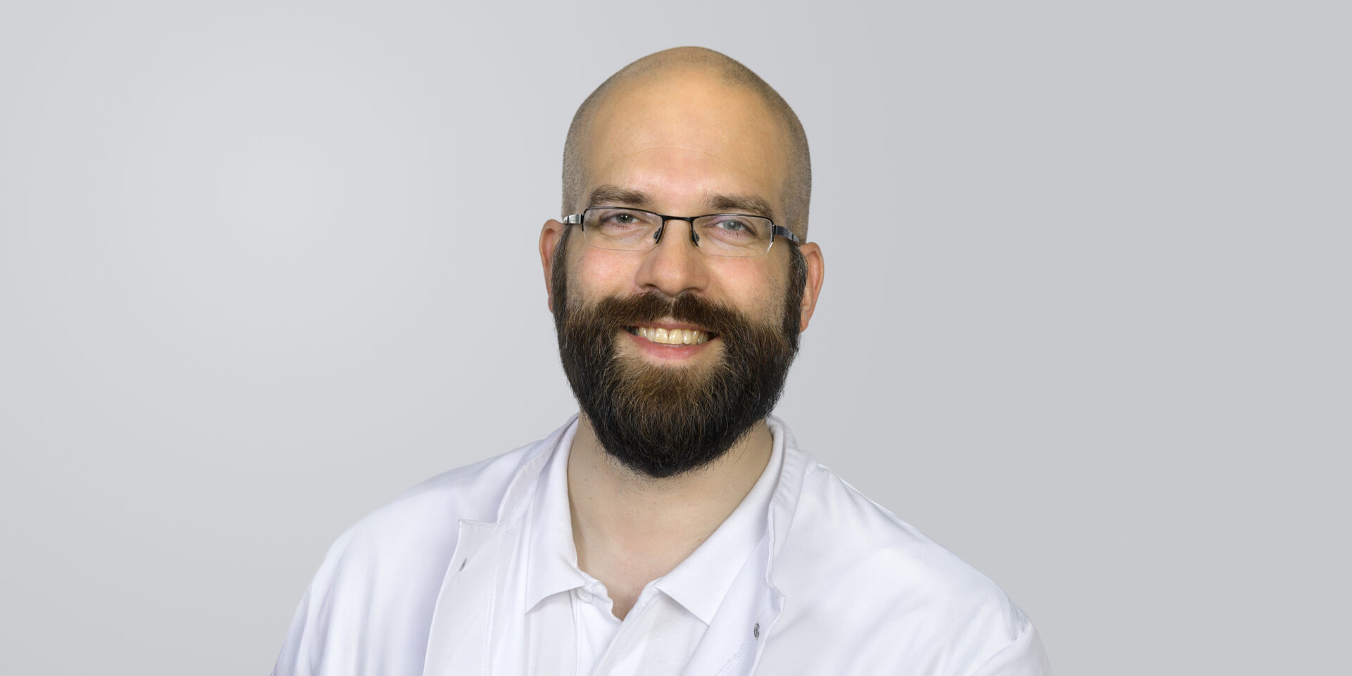 Lächelnder Mann mit Brille und Vollbart, trägt einen weißen Kittel vor neutralem Hintergrund.
