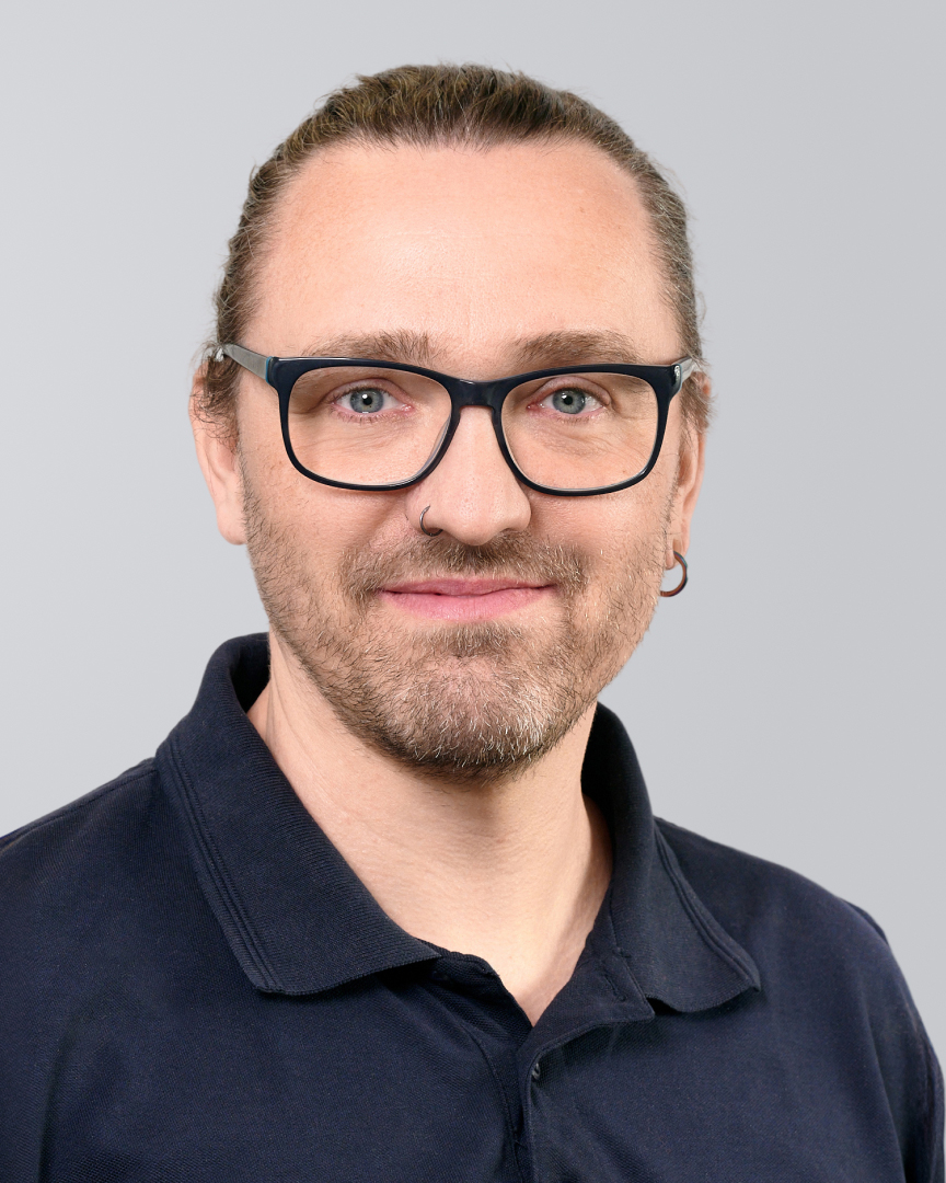 Lächelnder Mann mit Bart und Brille trägt ein schwarzes Polohemd vor einem neutralen Hintergrund.