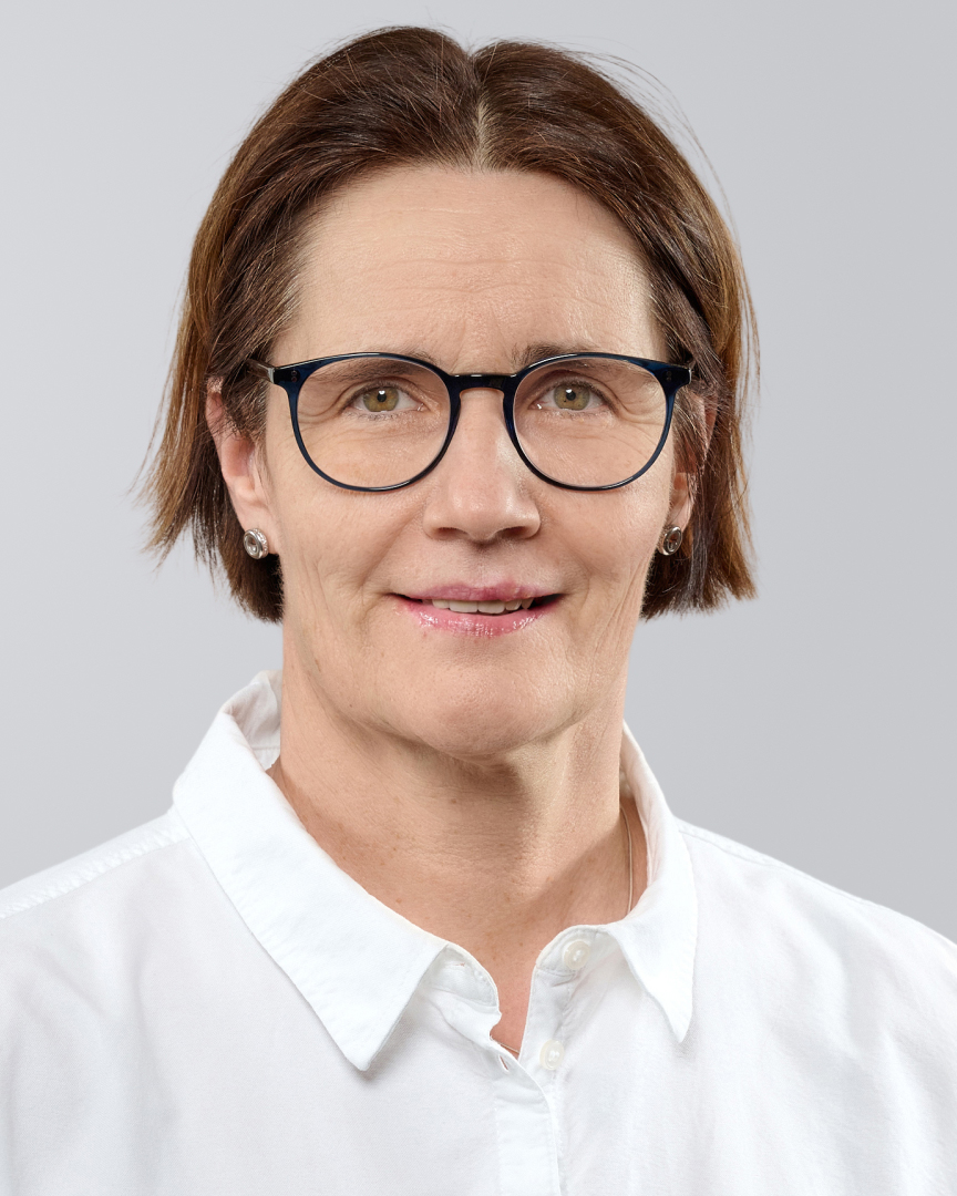 Porträt einer Frau mit kurzen, braunen Haaren, Brille und weißem Oberteil vor einem grauen Hintergrund.