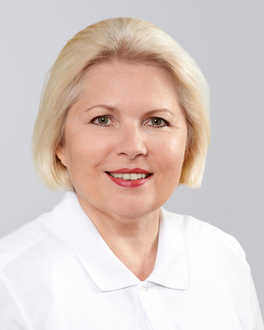 Porträt einer lächelnden Frau mit blonden, kinnlangen Haaren und weißer Bluse vor neutralem Hintergrund.