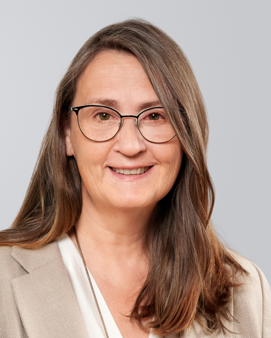 Ältere Frau mit langen, braunen Haaren, Brilleund beigefarbenem Blazer vor hellem Hintergrund.