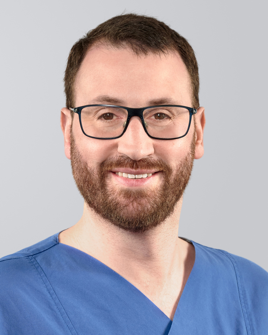 Mann mit Brille, Bart und blauer Arbeitskleidung lächelt vor einem grauen Hintergrund.