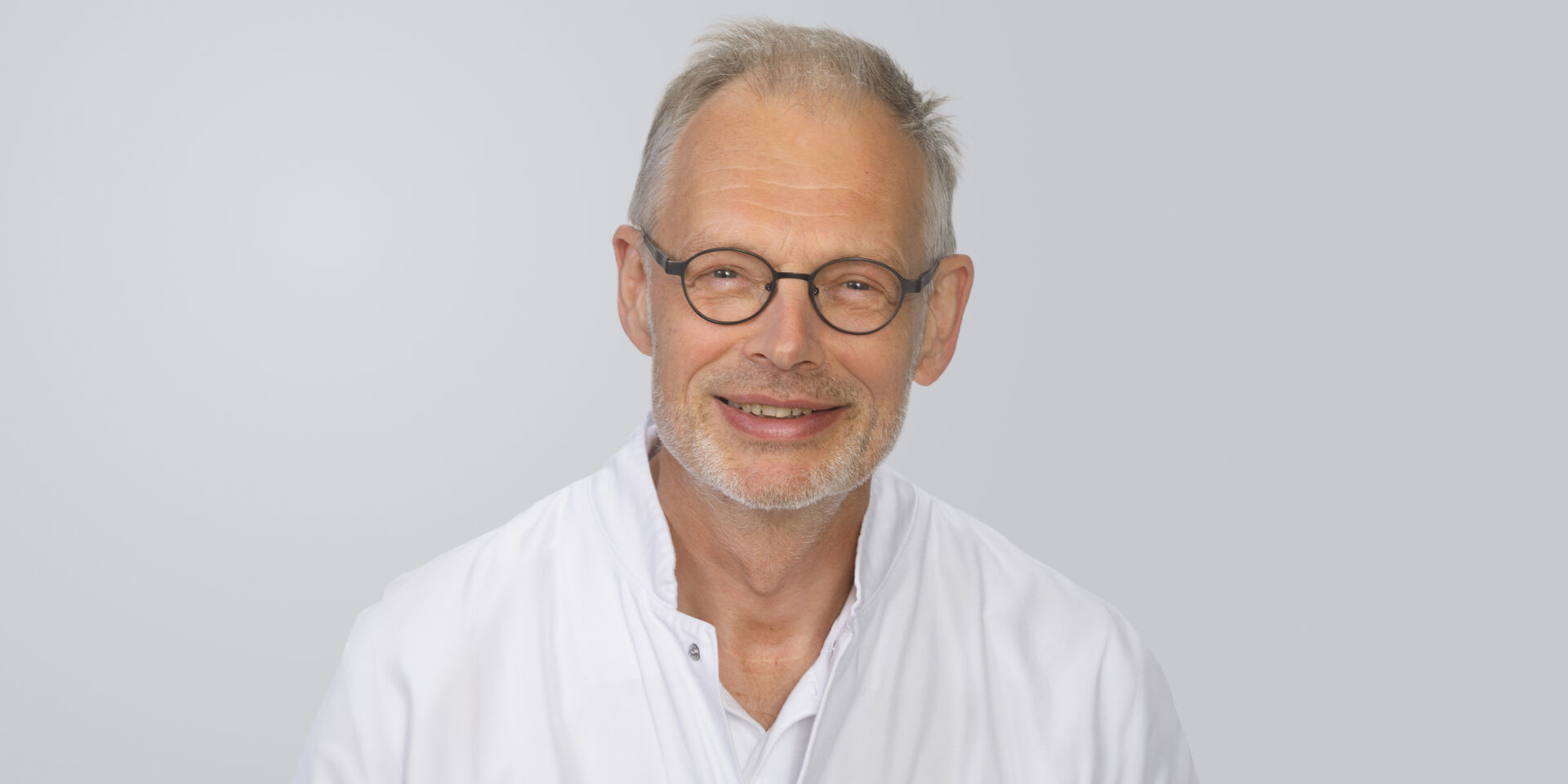 Älterer Mann mit Brille, grauen Haaren und weißem Kittel vor einem neutralen Hintergrund.