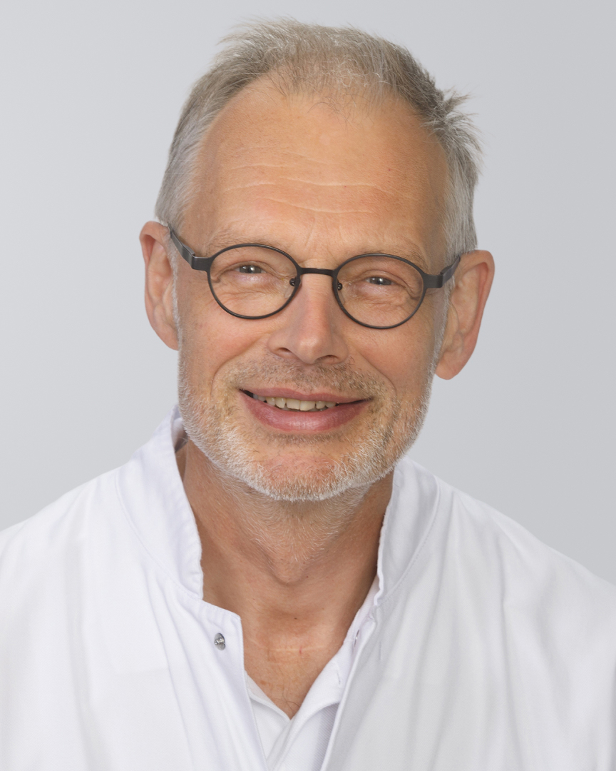 Älterer Mann mit Brille, grauen Haaren und weißem Kittel vor einem neutralen Hintergrund.