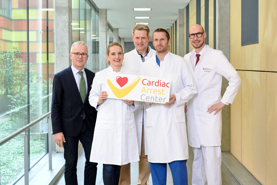 Eine Gruppe Menschen in Arztbekleidung steht in einem Flur. Eine junge Ärztin und ein junger Arzt halten ein Plaket mit der Aufschrift "Cardiac Arrest Center" in die Kamera.