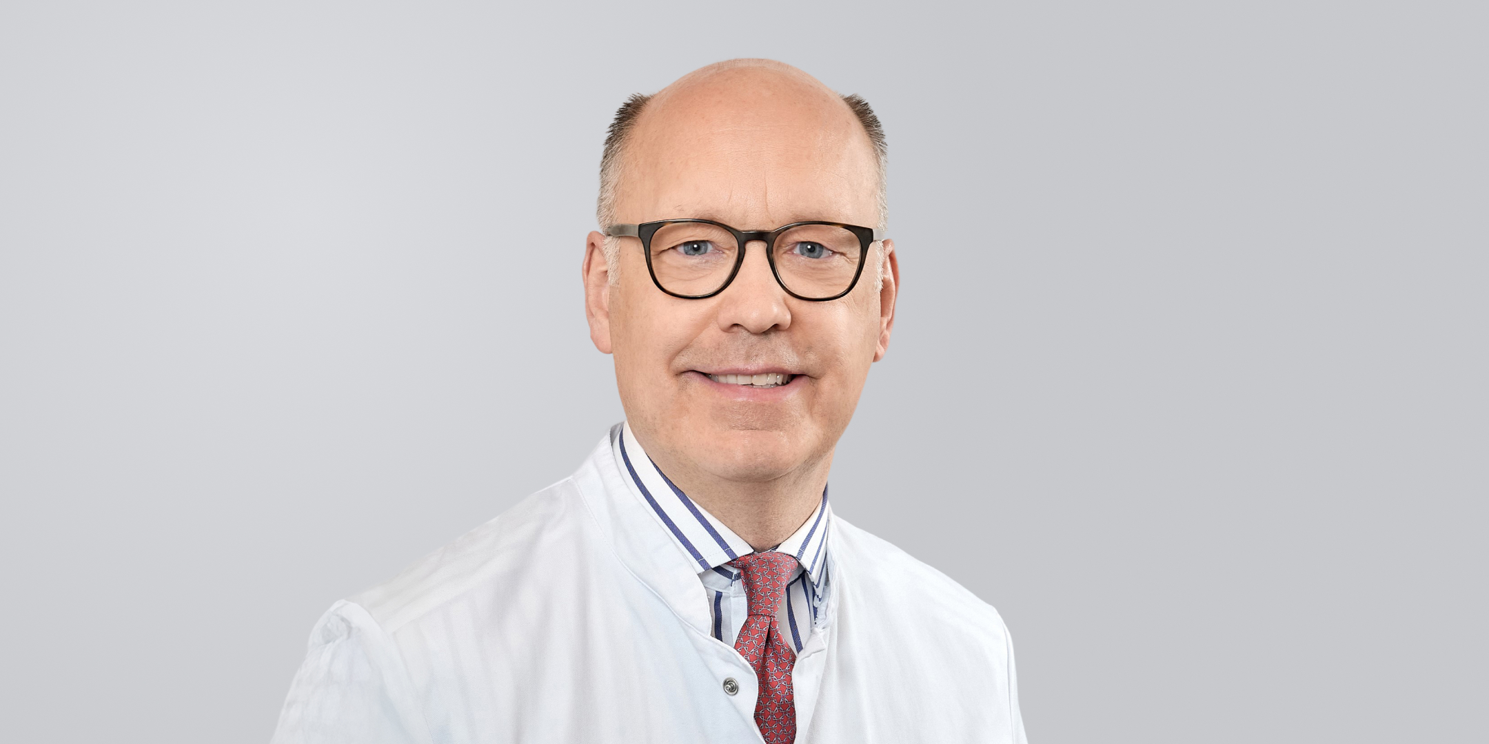 Ein lächelnder Arzt mit weißem Kittel, Brille und Krawatte vor einem grauen Hintergrund.