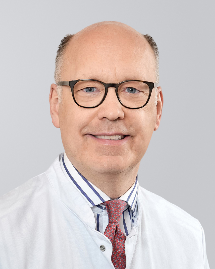 Ein lächelnder Arzt mit weißem Kittel, Brille und Krawatte vor einem grauen Hintergrund.