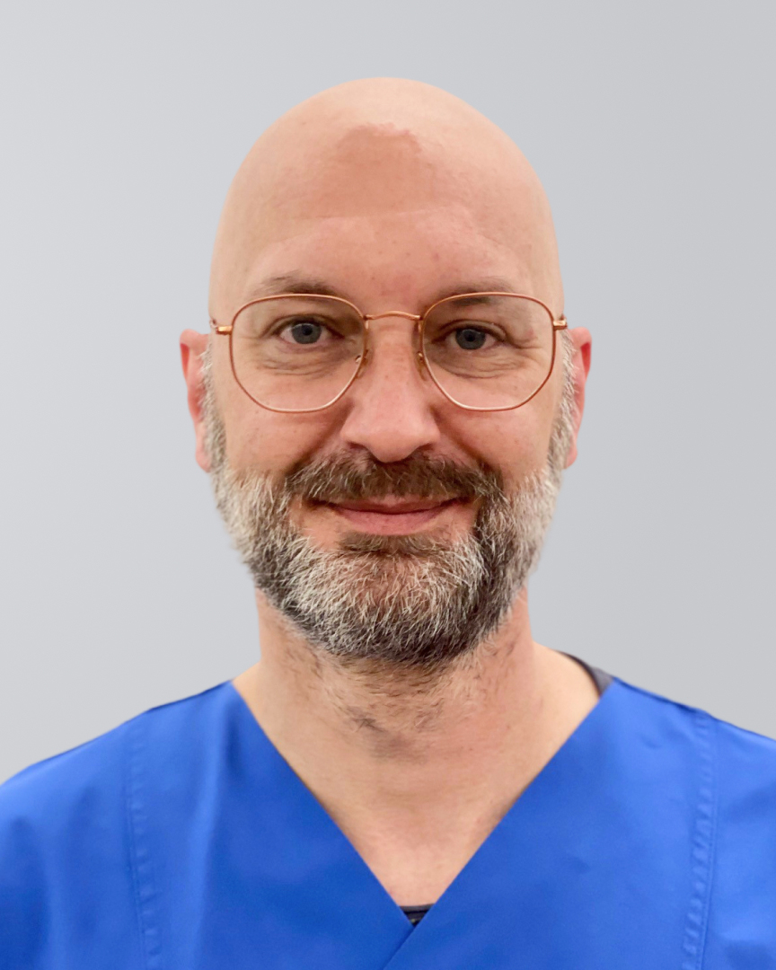 Mann mit Glatze, Bart, Brille und blauer Arbeitskleidung lächelt vor einem grauen Hintergrund.