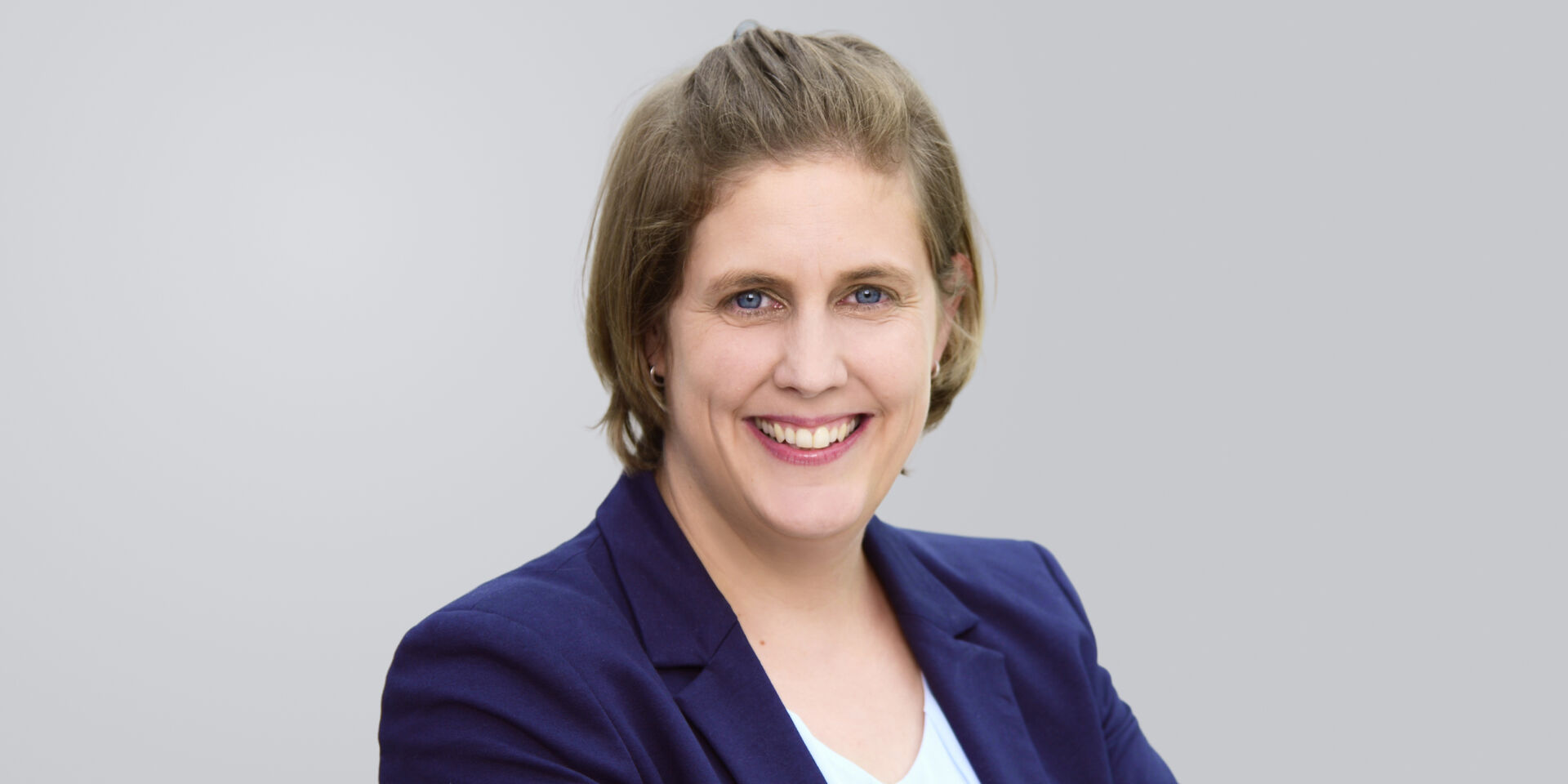 Lächelnde Frau mit kinnlangen Haaren und blauem Blazer vor einem grauen Hintergrund.